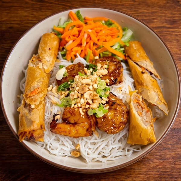 A bowl of Vietnamese vermicelli noodles with grilled shrimp, spring rolls, and fresh vegetables.