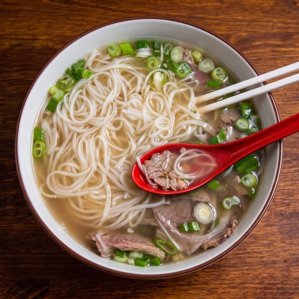 A bowl of pho with noodles, beef, and green onions, with chopsticks and a red spoon.